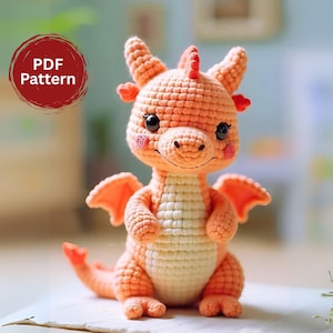 Pode incluir: Um dragão de crochê em tons de pêssego e creme. O dragão tem olhos grandes e pretos, asas pequenas e uma cauda longa. O texto "PDF Pattern" está em um círculo vermelho no canto superior esquerdo. É um item feito à mão.