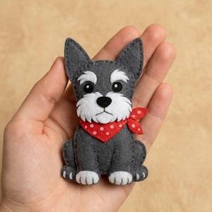 Puede incluir: Figura de perro de fieltro gris con detalles blancos alrededor del hocico y los ojos, y un pañuelo rojo con lunares. El perro está sentado erguido con las patas visibles. El fondo es de color beige liso.