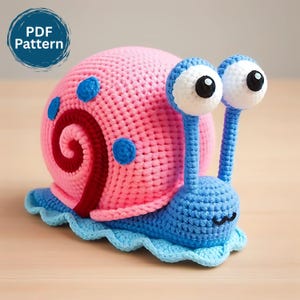 Puede incluir: Un caracol de ganchillo hecho a mano en rosa, azul y rojo. El caracol tiene ojos grandes y saltones y un diseño de caparazón en espiral. El texto "PDF Pattern" está en un círculo azul. El caracol está sobre una base azul claro.