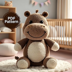 Op de afbeelding: Een gehaakt nijlpaard knuffel in bruin- en crèmetinten, met een lachend gezicht en kleine oren. Het nijlpaard heeft witte accenten op de voeten en buik. De afbeelding bevat de tekst "PDF Pattern" in een bruine cirkel.