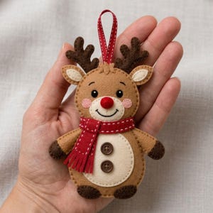 May include: A felt reindeer ornament featuring a tan body, dark brown antlers, and dark brown hooves. The reindeer has a bright red nose, pink cheeks, and a red scarf with white stitching. Two brown buttons are on the belly, and a red ribbon loop.
