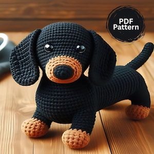 May include: A crocheted dachshund dog in black and tan. The dog has large, floppy ears, a long body, and short legs. The text "PDF Pattern" is in a black circle in the upper right corner. The dog is sitting on a wooden surface.