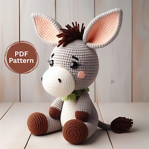 Puede incluir: Un peluche de burro tejido a ganchillo en tonos grises, marrones y blancos, con orejas grandes y un collar verde. El burro tiene una melena y cola marrones y esponjosas, y el texto "PDF Pattern" está en un círculo marrón.