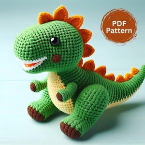 Może przedstawiać: Zielony szydełkowy dinozaur z pomarańczowymi kolcami i brązowymi detalami. Dinozaur ma uśmiechniętą twarz z białymi zębami i czarnymi oczami. Tekst "PDF Pattern" znajduje się w brązowym okręgu.