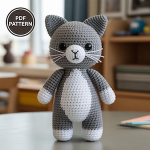 May include: A crocheted cat doll in grey and white, featuring black eyes and white whiskers. The cat has a white chest and paws. The image includes a circular "PDF PATTERN" graphic. The cat is standing on a surface.