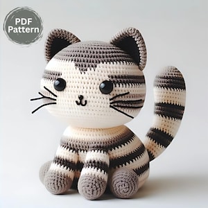 May include: A handmade crochet cat toy in cream, grey and dark grey stripes. The cat has large black eyes, whiskers, and a small nose. The image includes a circular graphic that says "PDF Pattern".