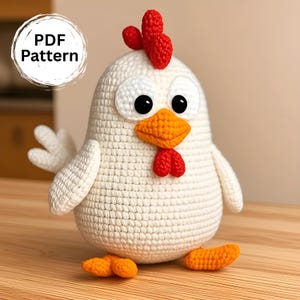 Peut inclure: Un poulet blanc crocheté avec une crête rouge, une barbichette et un bec et des pieds orange. Le poulet a de grands yeux noirs et des contours d'yeux blancs. Le texte "PDF Pattern" est dans un cercle blanc en haut à gauche. Le poulet est posé sur une surface en bois.