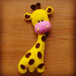 Könnte beinhalten: Eine handgefertigte Giraffe aus gelbem Filz mit braunen Flecken, Ohren und Hörnern. Die Giraffe hat eine rosa Nase und kleine schwarze Augen. Dieses Plüschtier ist ein niedliches und kuscheliges Produkt.