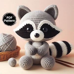 Könnte beinhalten: Ein gehäkeltes Waschbär-Plüschtier mit grauen, weißen und schwarzen Details. Der Waschbär hat große schwarze Augen, eine weiße Schnauze und einen gestreiften Schwanz. Ein Knäuel grauer Wolle und ein Korb befinden sich im Hintergrund. Das Bild enthält ein "PDF Pattern"-Etikett.
