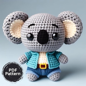 Puede incluir: Koala de ganchillo gris, con una chaqueta y pantalones cortos vaqueros azules con cinturón marrón. El koala tiene orejas grandes y redondas con el interior color crema, ojos negros y nariz negra. Un gráfico circular con el texto "PDF Pattern" está en la esquina inferior izquierda.