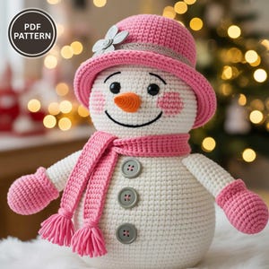 May include: A crocheted snowman wearing a pink hat, scarf, and mittens. The snowman has a carrot nose, black eyes, and a smiling mouth. The snowman has three gray buttons down the front. The image includes the text "PDF PATTERN".