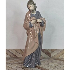 May include: A ceramic figurine of a man in a long robe, with a beard and downward gaze. The robe is a combination of grey and peach tones. The figurine is standing on a wooden floor.