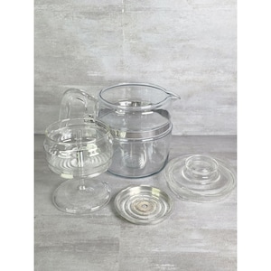 Puede incluir: Un juego de cafetera de goteo de vidrio transparente que incluye una jarra grande con asa, un conjunto de cesta de vidrio con vástago central, una placa de filtro metálica circular con pequeñas perforaciones y una tapa de vidrio a juego.