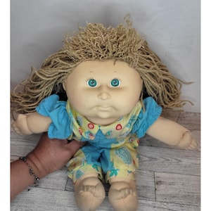 Könnte beinhalten: Vintage Puppe mit weichem Stoffkörper und Vinylgesicht. Die Puppe hat lockiges, hellbraunes Haar und große, grüne Augen. Sie trägt ein blau-gelbes Outfit mit Paisley-Muster. Die Arme und Beine der Puppe sind beweglich.