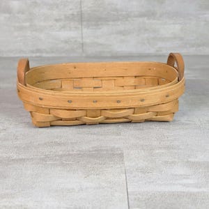 Puede incluir: Una cesta ovalada de madera tejida con un acabado de color tostado claro. La cesta tiene un borde reforzado con remaches metálicos visibles y dos asas de madera resistentes unidas a los lados. La estructura consta de tiras de madera planas.