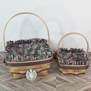 Könnte beinhalten: Zwei Osterkörbe mit Holzgriffen. Die Körbe sind mit hellbraunen, gelben und rosa Streifen gewebt. Die Oberseiten sind mit einem floralen Stoff mit Ostereiern und Blumen gefüttert. Ein Anhänger mit dem Wort "Ostern" hängt am größeren Korb.