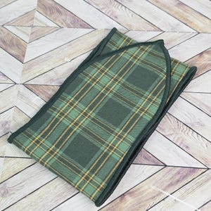 May include: A folded green and gold plaid pocket square with a dark green border. The fabric has a classic tartan pattern. The pocket square is displayed on a light wood surface.
