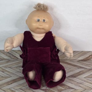 Könnte beinhalten: Eine Vintage Cabbage Patch Kid Puppe mit kahlem Kopf und einem Büschel hellbrauner Haare. Die Puppe trägt einen weinroten Cord-Overall mit einer Tasche und dekorativen Bindungen. Die Puppe hat blaue Augen und einen neutralen Gesichtsausdruck.