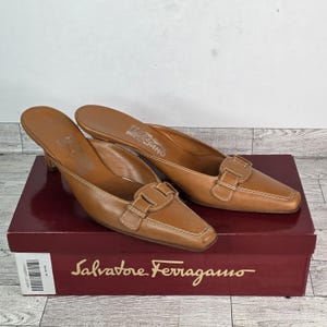 Puede incluir: Un par de mules Salvatore Ferragamo de cuero marrón con tacón pequeño y punta cuadrada. Los zapatos tienen un detalle de hebilla y costuras blancas. Se exhiben en una caja roja Salvatore Ferragamo.