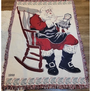 May include: A woven throw blanket featuring Santa Claus in a rocking chair, holding a doll. Santa is dressed in a red suit with black boots. The blanket has a cream background with a holly and berry border and the year "1992".