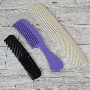 May include: Three combs of different colors and sizes are displayed on a wooden surface. A black comb, a purple comb with a handle, and a beige comb are visible. The beige comb has the word "Unbreakable" printed on it.