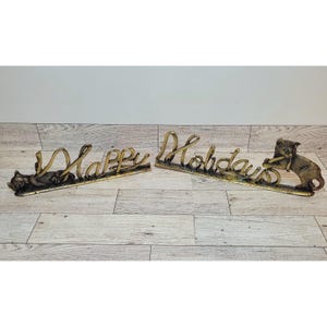 May include: Two metal holiday signs with the words "Happy Holidays". One sign has a cat lying down, and the other has a cat playing with the word "Holidays". The signs are gold and brown.