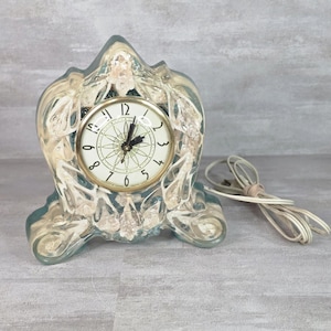 May include: A translucent, light blue resin mantel clock with an ornate, molded frame featuring embedded floral accents. The clock face is circular with a gold-tone rim, black Arabic numerals, and a central geometric star pattern. Includes a white cord.