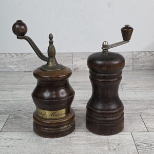 Puede incluir: Dos molinillos de pimienta antiguos de madera. Uno tiene un mango de latón y una etiqueta que dice "Verde Muskat". El otro es un diseño simple de madera oscura. Ambos molinillos tienen una estética clásica y vintage.