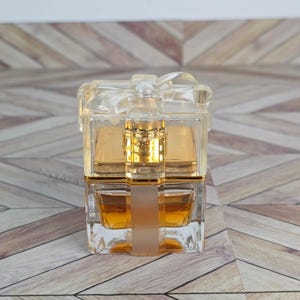 May include: A clear, rectangular perfume bottle with a bow-shaped top. The bottle contains a golden-colored liquid. The design is a gift box shape, with a frosted vertical band. The bottle sits on a wooden surface.