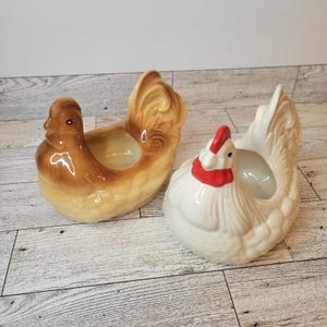 May include: Two ceramic hen-shaped planters. One is brown with a glossy finish, and the other is white with a red comb and wattle. Both have a hollowed-out center for planting small flowers or herbs. The planters are decorative.