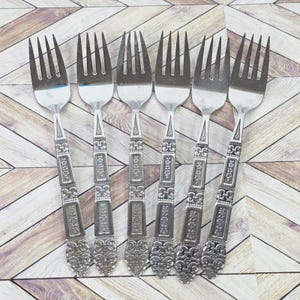 May include: Six silver forks with detailed, ornate handles. The forks have a classic design with a decorative pattern on the handles and tines. The forks are arranged on a light-coloured wooden surface.