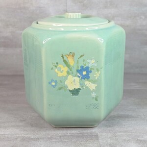 Pode incluir: Um recipiente de cerâmica em verde menta claro com formato hexagonal e tampa a condizer. A parte frontal apresenta um design floral pintado com flores amarelas, azuis e brancas e folhas verdes. A superfície tem um acabamento liso e brilhante.