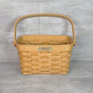 Puede incluir: Una cesta rectangular de madera tejida con un asa arqueada. La cesta presenta un acabado natural en tono bronceado claro y una placa de metal color latón en la parte frontal con el texto Longaberger grabado en una fuente de estilo cursiva.