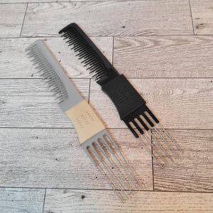 Könnte beinhalten: Zwei Haarkämme auf einer Holzoberfläche. Ein Kamm ist grau und beige mit Metallzinken, der andere ist schwarz mit Metallzinken. Beide Kämme haben einen Griff und Zähne zum Frisieren.