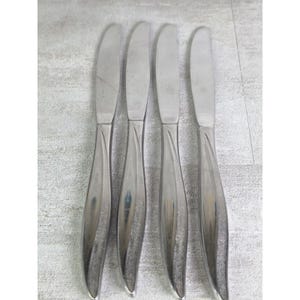 May include: A set of four silver-toned stainless steel dinner knives arranged vertically. Each knife features a smooth, polished blade and a handle with a textured, matte finish along the center, tapering toward the base of the handle.