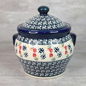 Puede incluir: Un azucarero de cerámica con tapa, que presenta una base de color crema decorada con un patrón repetitivo de pequeñas cerezas rojas, ramitas de hojas azul oscuro y bordes festoneados azul oscuro alrededor del borde, la base y la tapa.