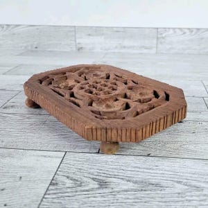May include: A rectangular, brown wooden trivet with carved floral and geometric designs. The trivet is raised on four small, rounded feet. The wood has a natural, warm tone, and the carving is detailed.