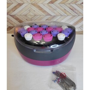 May include: A hair roller set with a clear plastic lid, featuring a black and magenta base. The set includes various colored rollers: white, pink, and purple. A small bag of silver hair pins is also included.