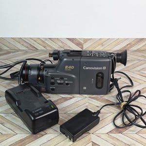 Puede incluir: Videocámara Canon Canovision 8 gris oscuro con lente y cable de alimentación. La videocámara tiene el número de modelo E40. También se incluye un cargador de batería Canon CA-100 y un adaptador de corriente negro.