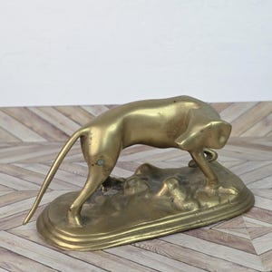 May include: A vintage brass sculpture of a dog in a hunting pose. The dog is depicted with its head down, tail extended, and front leg bent. The sculpture is mounted on a textured base with an oval shape.
