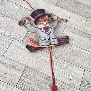 May include: A wooden pull-along toy in the shape of a cat. The cat is dressed in a top hat, a light blue coat, and red striped trousers. It holds a candy cane and a bird. A red string with a red ball hangs.