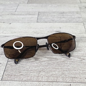 May include: Brown-tinted sunglasses with a dark metal frame. The lenses are rectangular and the frame has a thin, wire-like design. The temples are black and feature a small logo. The sunglasses are resting on a light gray wooden surface.