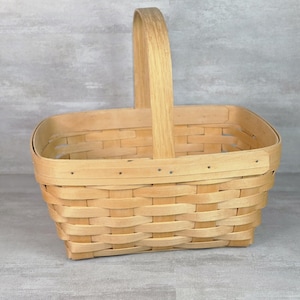 May include: A rectangular woven wood basket featuring a light tan natural finish. The basket has a single tall arched handle attached to the centre of the rim and is constructed with horizontal and vertical strips of wood secured by small metal nails.