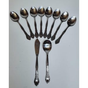 Könnte beinhalten: Eine Sammlung von Edelstahlbesteck, darunter acht Löffel, ein Buttermesser und ein Servierlöffel. Das Besteck hat ein dekoratives Griffdesign und ist auf einer weißen Oberfläche angeordnet.