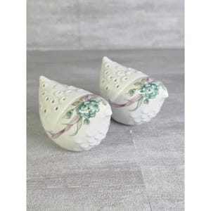 May include: A pair of white ceramic bird-shaped shakers featuring a delicate painted design of teal roses and light purple ribbons. The upper sections of the birds have small circular perforations, and the lower bodies have a textured scale pattern.