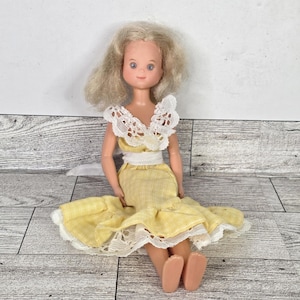 May include: A vintage doll with blonde hair and blue eyes, wearing a yellow dress with white lace trim. The doll's dress has a white belt and a ruffled skirt. The doll is sitting down.