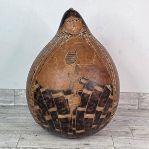 Può includere: Un grande vaso a forma di zucca marrone con disegni incisi. Il vaso presenta una figura centrale che tiene uno strumento, con motivi geometrici alla base. La parte superiore ha un disegno di volto. La zucca è di colore marrone naturale.