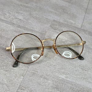 May include: Round eyeglasses featuring thin gold metal temples and a tortoiseshell pattern rim around the clear lenses. The bridge and hinges are constructed from gold-toned metal, and the temple tips are finished in a dark, solid material.