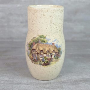 Puede incluir: Un jarrón cilíndrico de cerámica con un acabado en color crema moteado. La parte frontal presenta una ilustración detallada y colorida de una casa con techo de paja rodeada de árboles verdes y flores vibrantes bajo un cielo azul.
