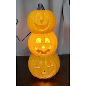 May include: A stacked Halloween pumpkin decoration with three carved, smiling faces. The pumpkins are illuminated from within, casting a warm orange glow. The top pumpkin has a dark stem. A black power cord is visible.